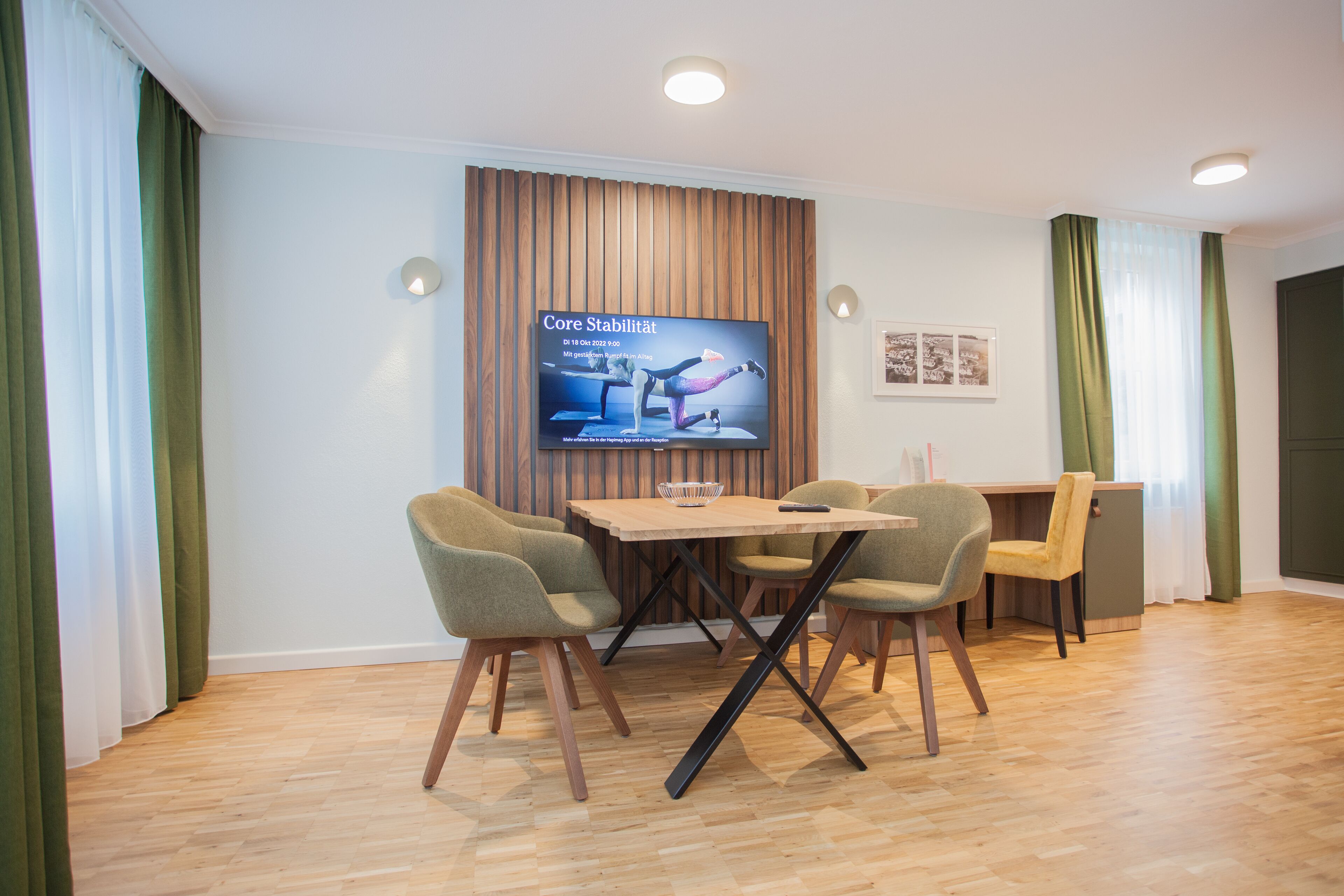 A modern conference room with a wooden table, four chairs, a TV screen displaying a yoga class, and a wooden wall panel.
