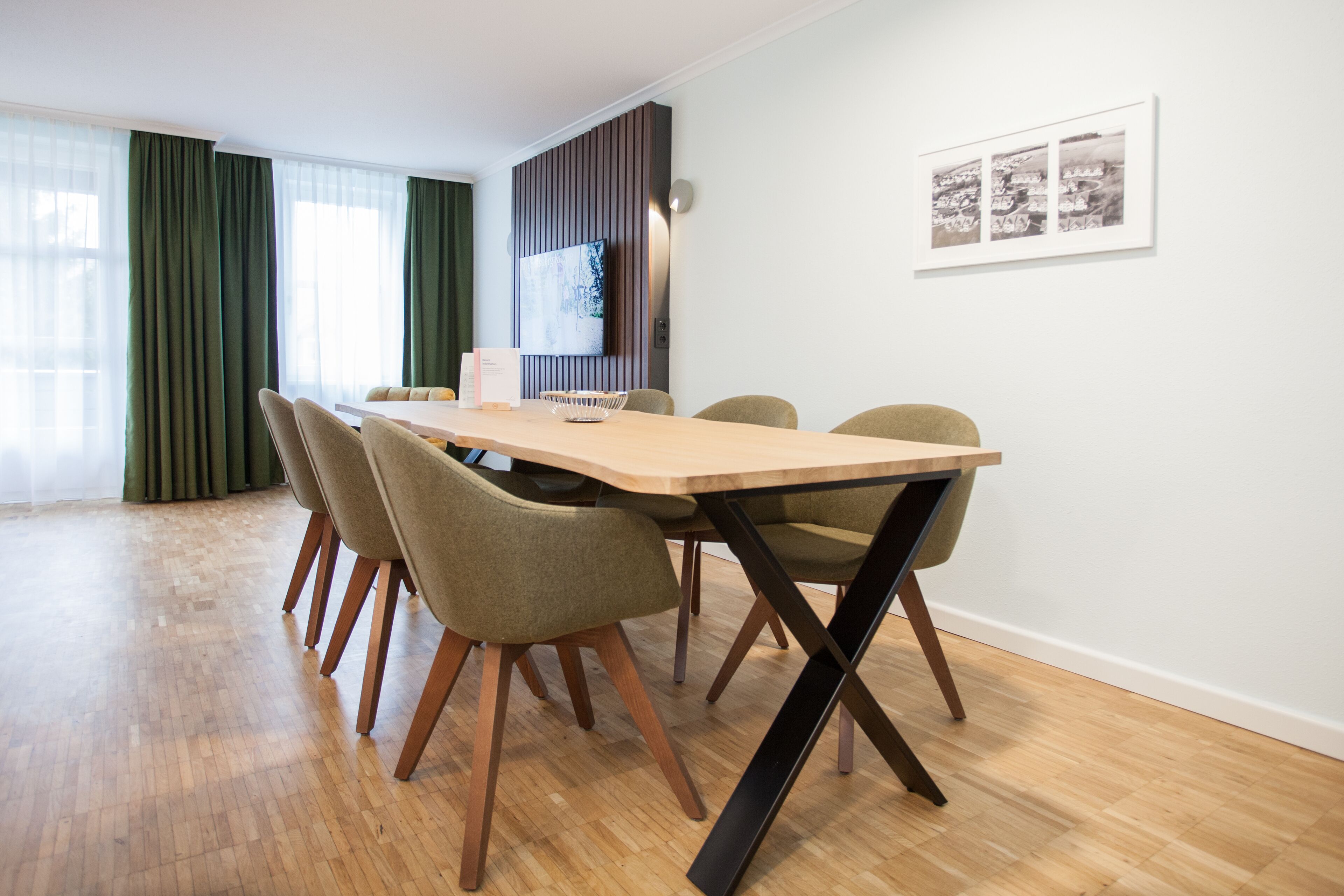 A modern dining area with a wooden table and four chairs, a flat-screen TV mounted on the wall, and a framed picture above the table.