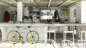 A modern outdoor bar area with a bicycle, white bar stools, and a view of palm trees. - Hotel 28 (Santorini)