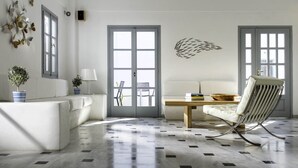 A modern living room with a white sofa, a dining table, and a striped chair. There are two large windows with shutters, a decorative wall piece, and a potted plant. - Hotel 28 (Santorini)