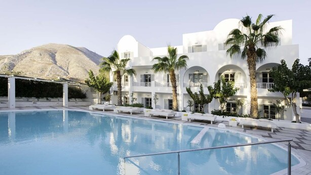 A poolside area with a clear blue pool, white buildings with balconies, and palm trees. - Hotel 28 (Santorini)
