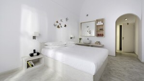 A minimalist bedroom with a large bed, bedside tables, a wall-mounted mirror, and a small shelf with toiletries. - Hotel 28 (Santorini)