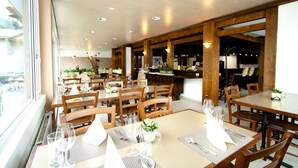 A restaurant with wooden tables and chairs, a bar area, and large windows.