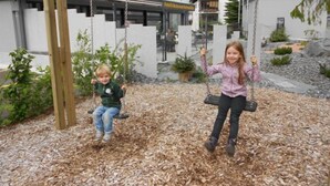 Two children on swings in a playground area with a modern building in the background.