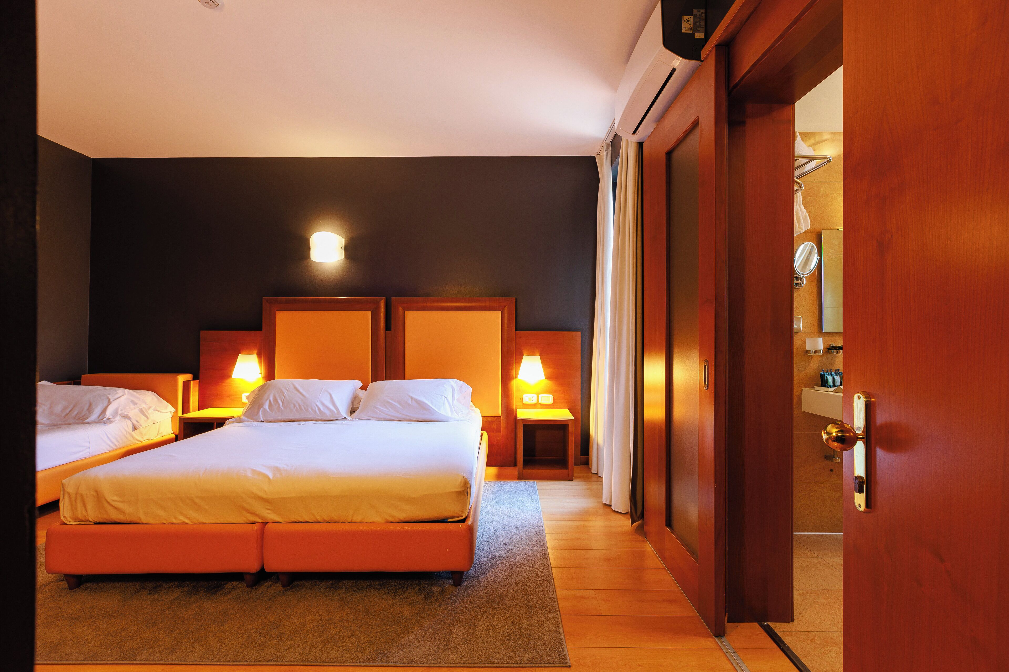 Habitación de hotel con dos camas, suelo de madera y baño visible a través de una puerta abierta.