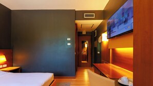 A hotel room with a bed, a desk, a chair, a TV mounted on the wall, and a bathroom visible in the background.