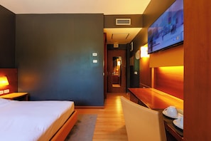 A hotel room with a bed, a desk, a chair, a TV mounted on the wall, and a bathroom visible in the background.