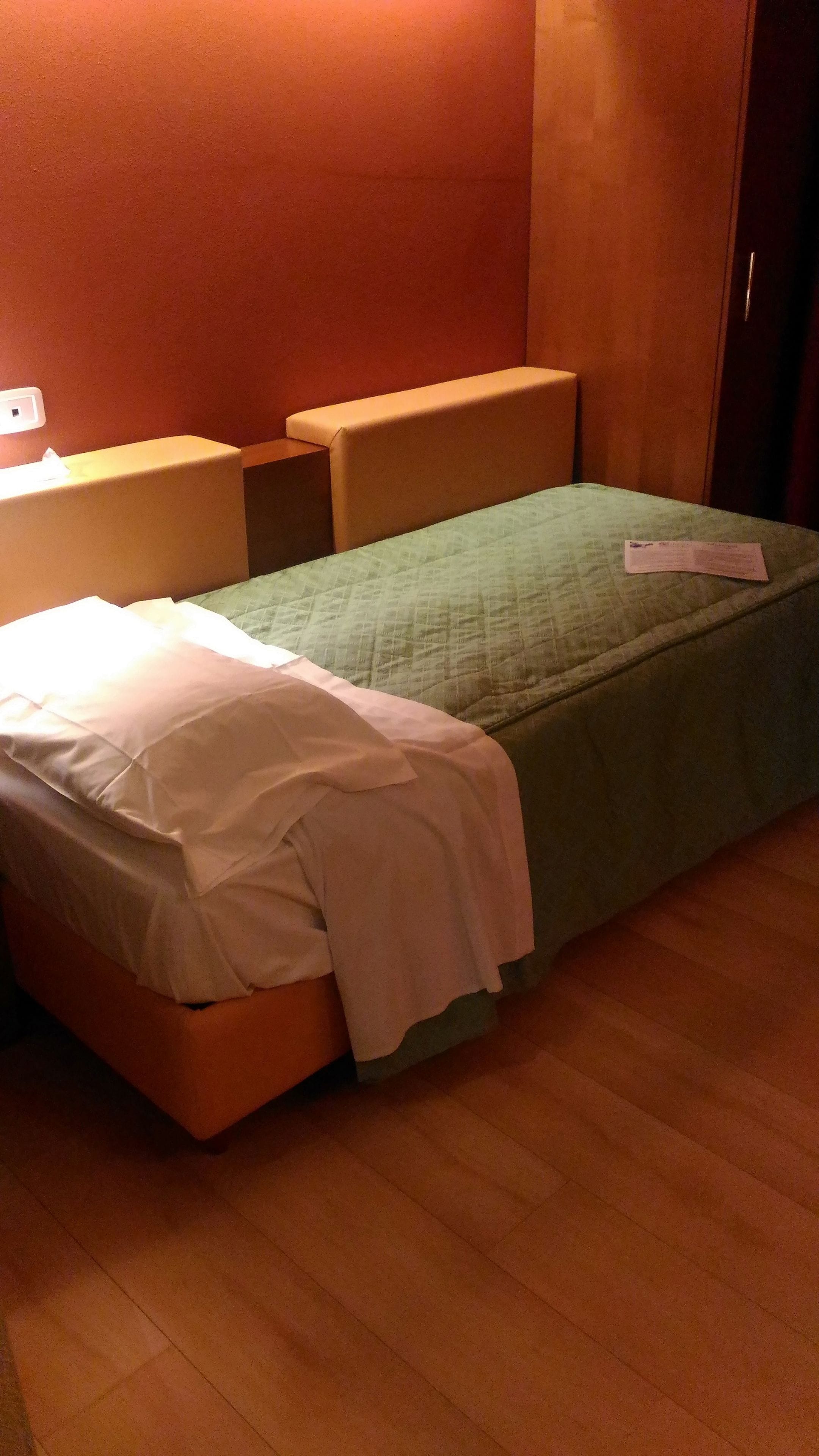 A small, single-bed room with a green bedspread and a white pillow. There is a wooden nightstand with a lamp on it. The room has wooden flooring and a red wall.