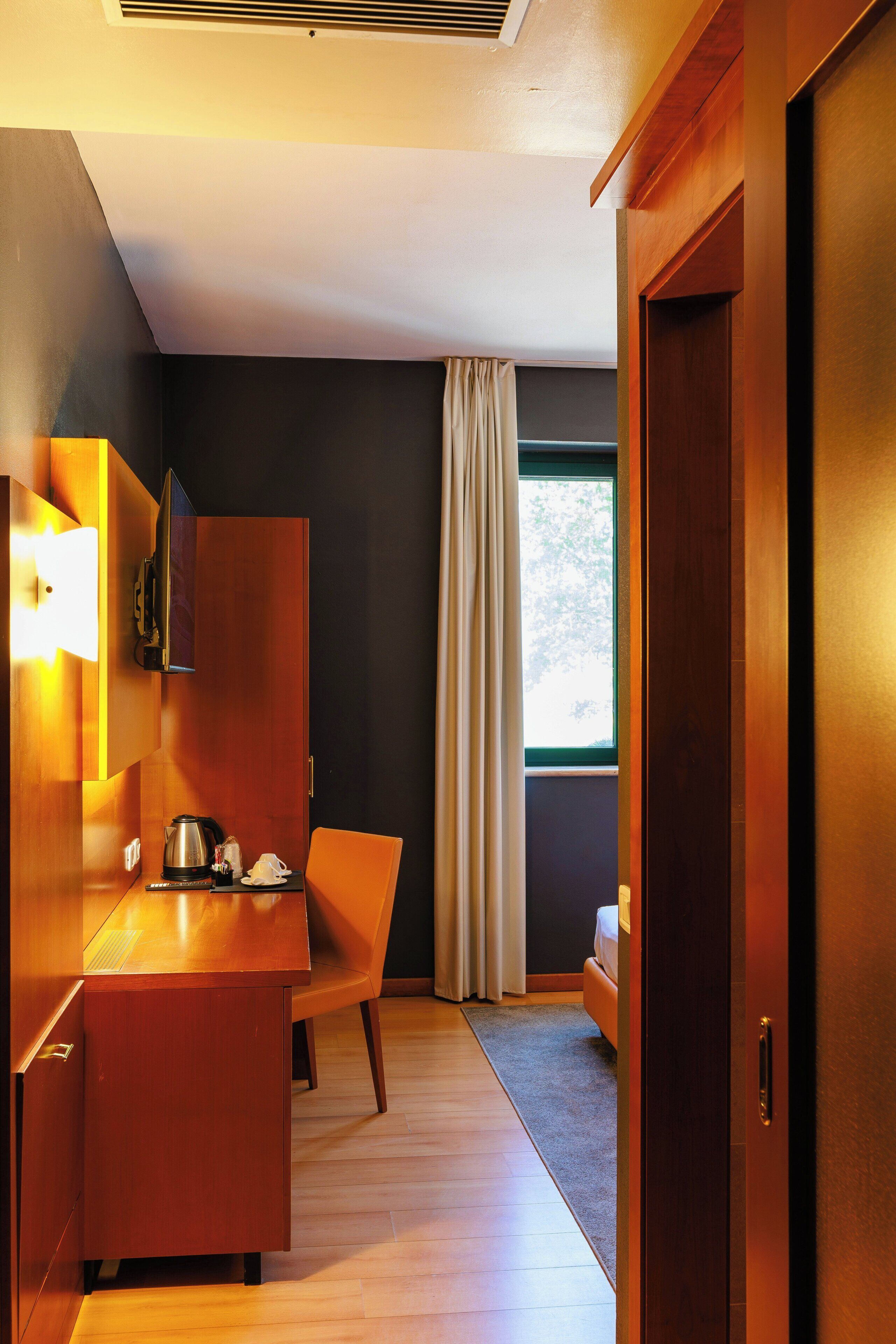 Habitación de hotel con escritorio de madera, una silla, una cama, ventana con cortinas y una puerta.