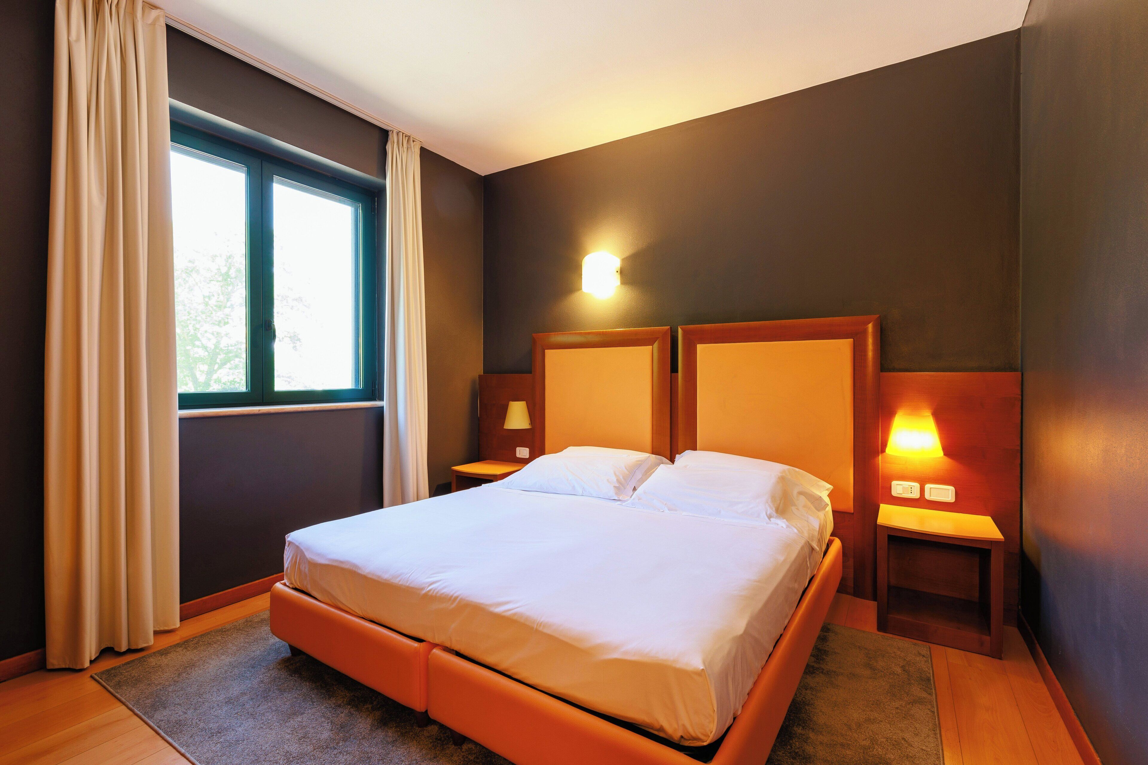A bedroom with a bed, a window with curtains, a nightstand with a lamp, and a wall-mounted light.