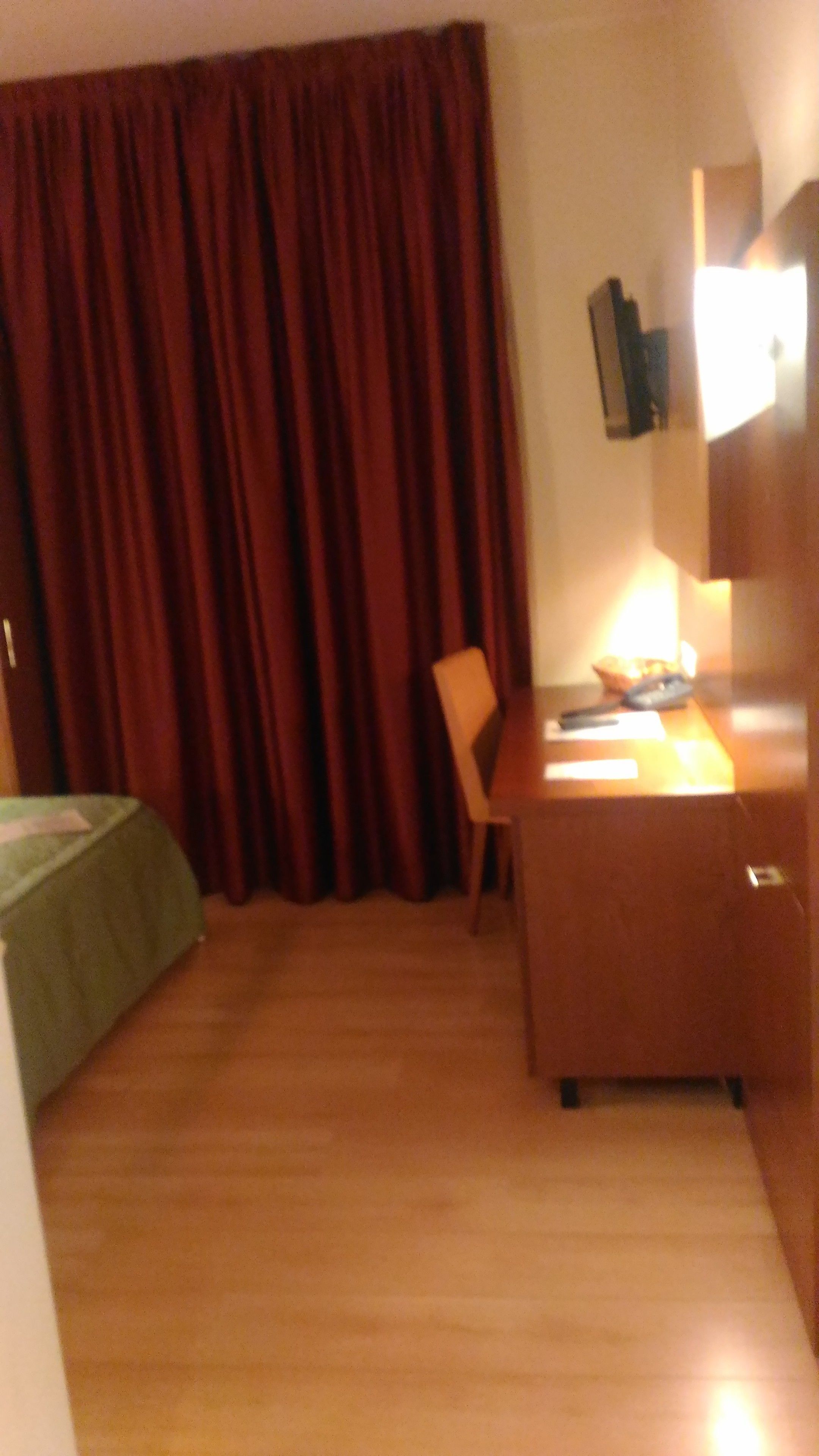 Habitación de hotel con un escritorio de madera, una silla, una cama con colcha verde, un ventanal con cortinas y un televisor de pared.