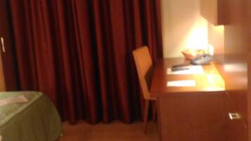 Habitación de hotel con un escritorio de madera, una silla, una cama con colcha verde, un ventanal con cortinas y un televisor de pared.