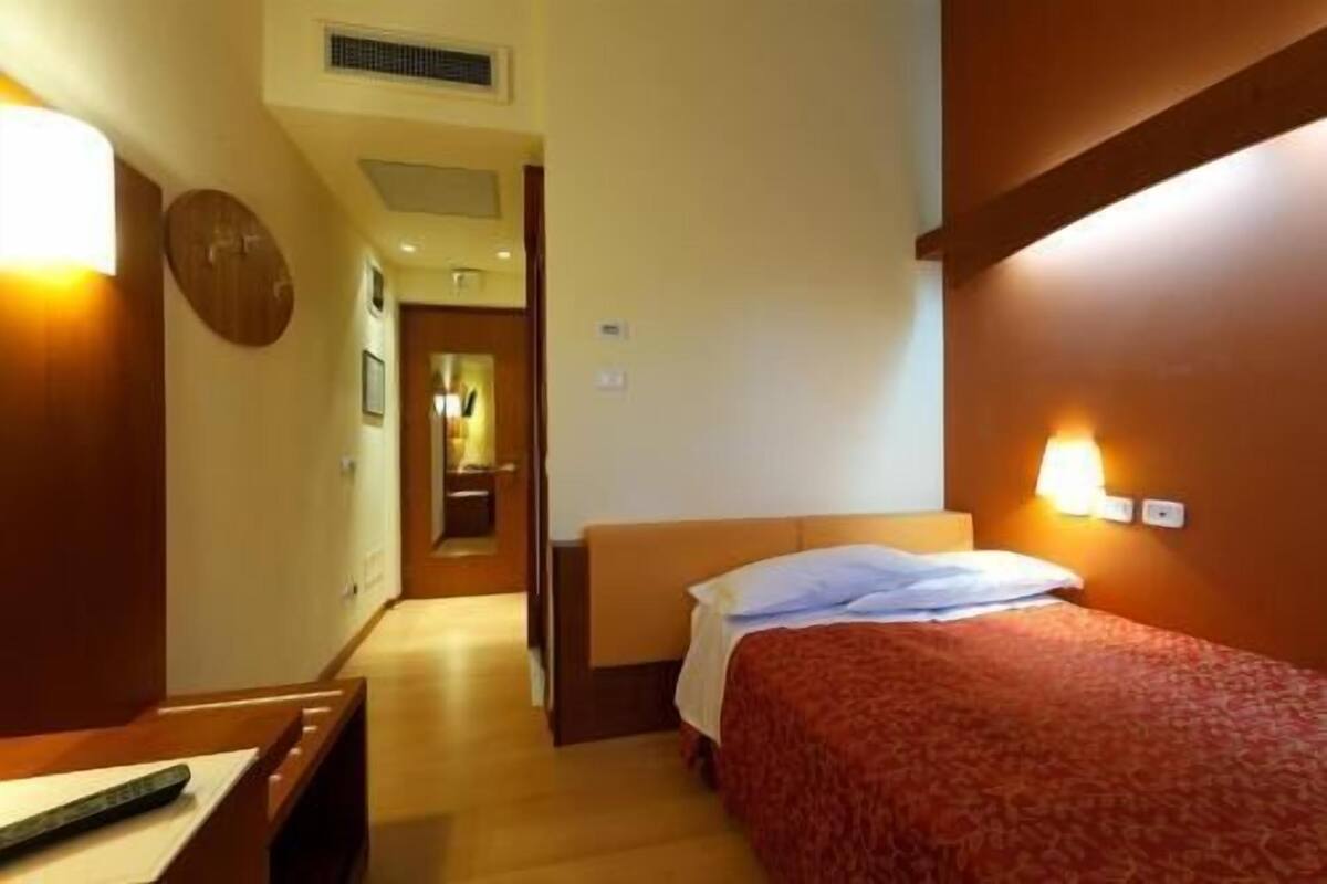 Een hotelkamer met een bed, een bureau met stoel, een nachtkastje, een wandlamp en een deur die naar een andere kamer leidt.