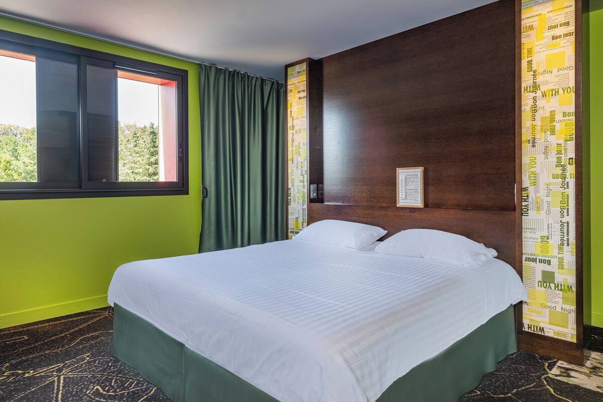 Une chambre d’hôtel avec un lit, des murs verts, une fenêtre avec des rideaux et une tête de lit en bois.