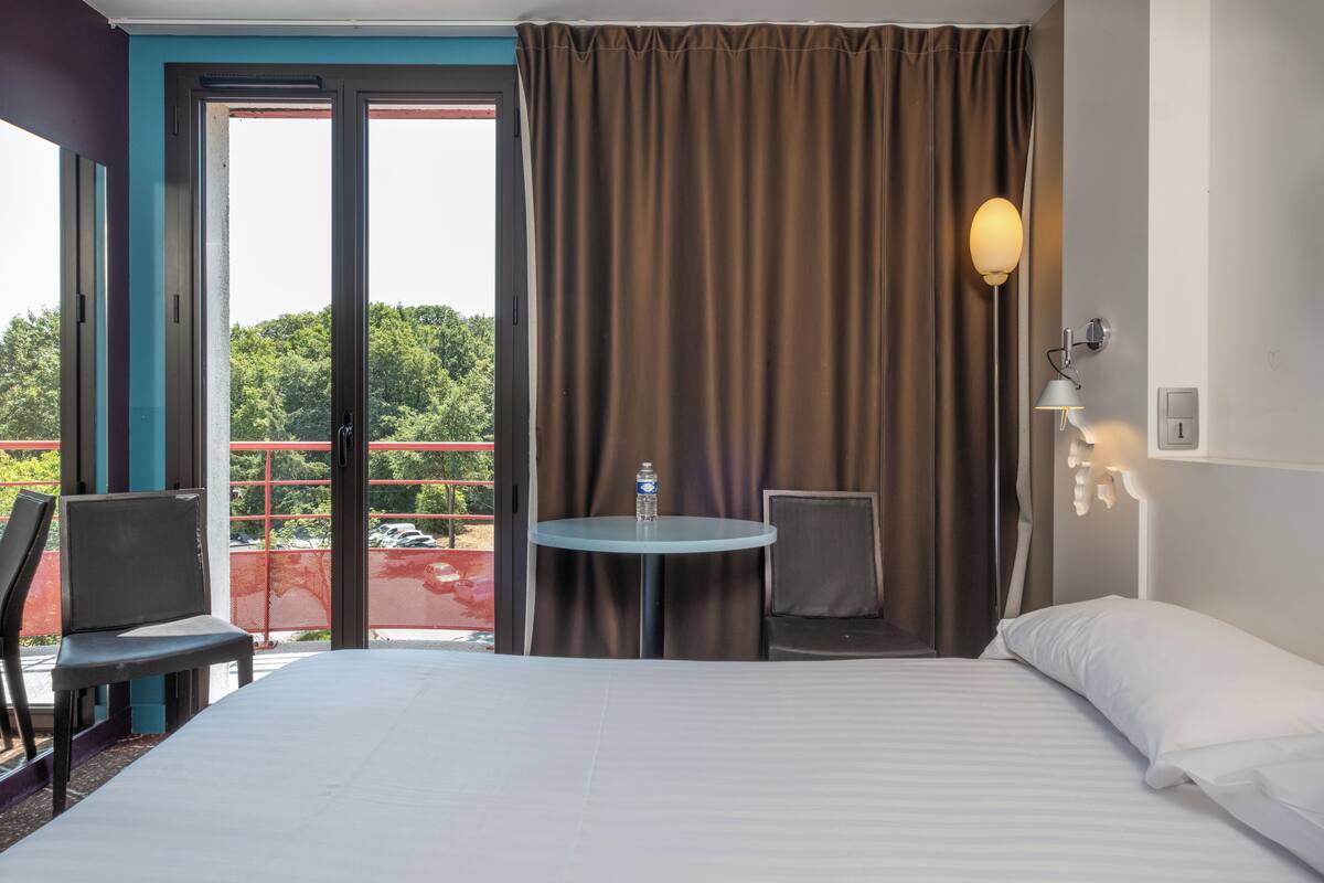 Une chambre d’hôtel avec un lit, une table ronde entourée de chaises, une bouteille d’eau sur la table et une vue sur l’extérieur par une porte coulissante.