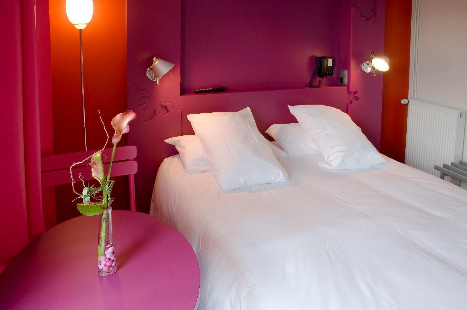 Habitación de hotel con una cama, almohadas blancas, una silla rosa, un florero con flores y un teléfono montado en la pared.