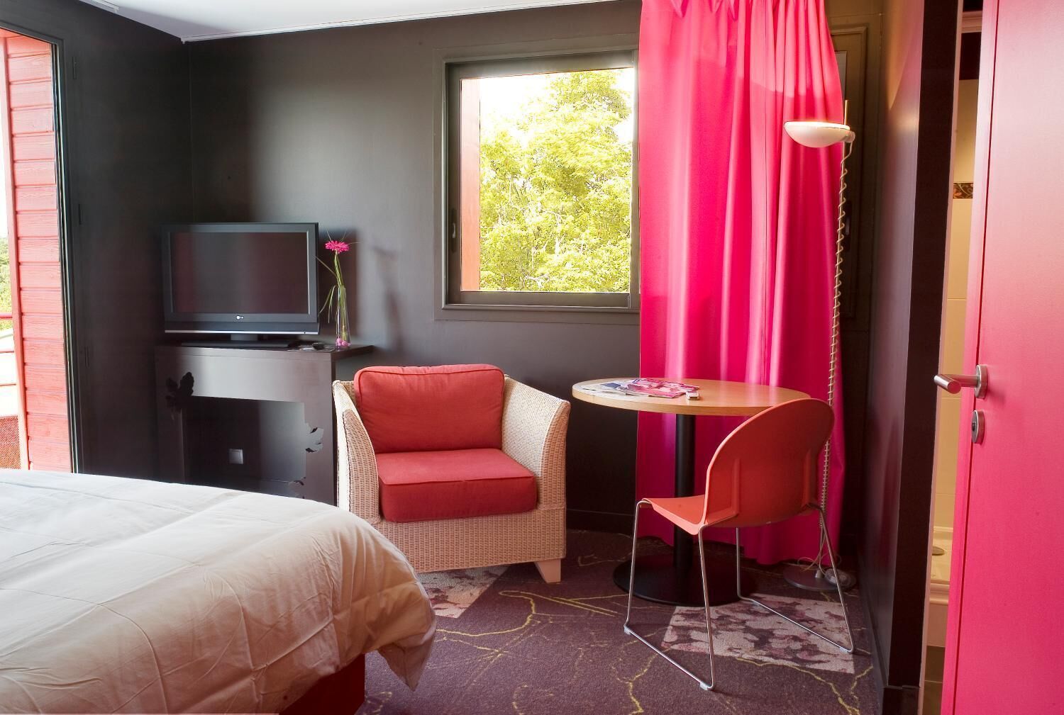 A hotel room with a bed, a television, a red chair, a small table, and a window with curtains.