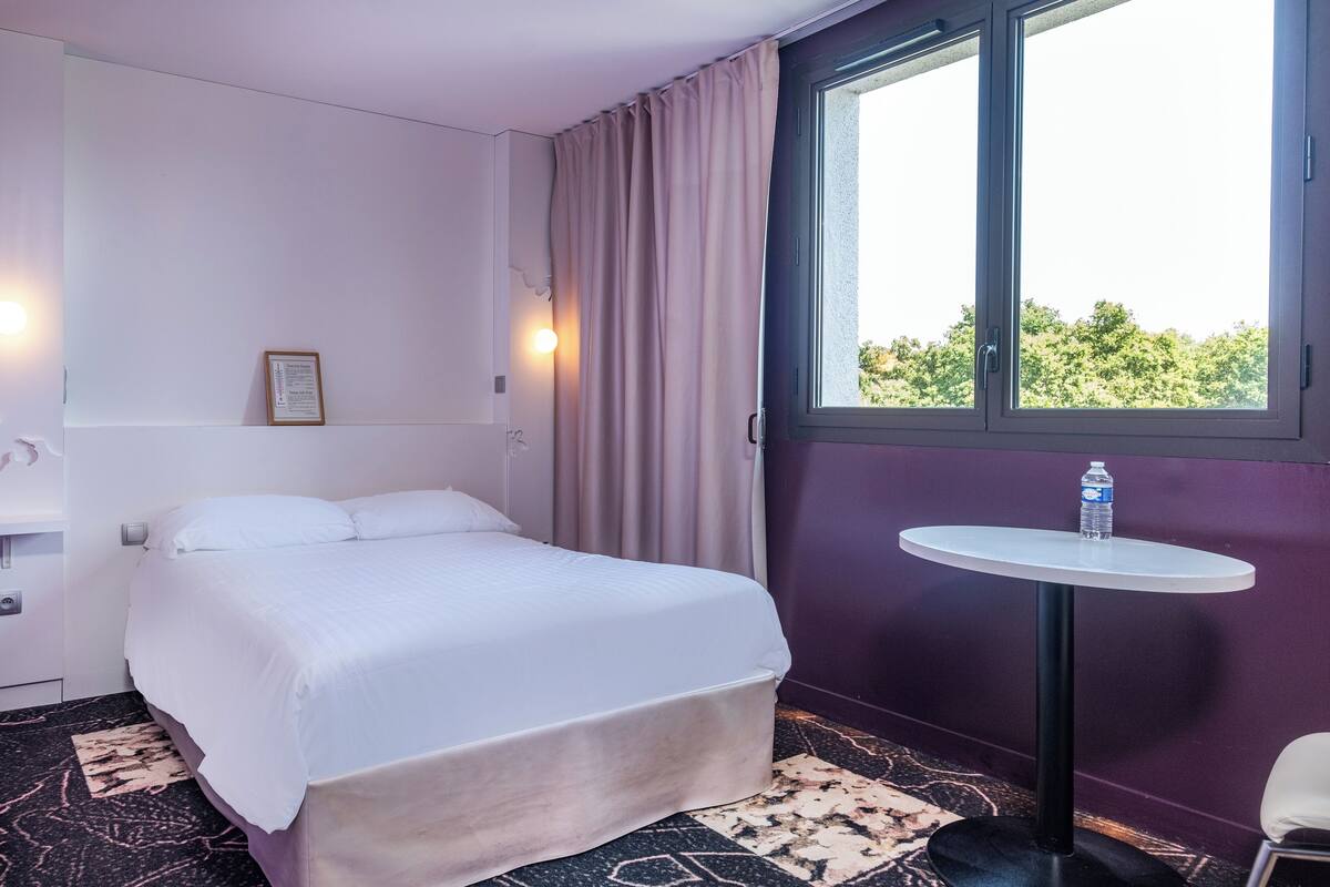 Une chambre d’hôtel avec un lit, une petite table ronde sur laquelle se trouve une bouteille d’eau, une fenêtre avec des rideaux et une lampe fixée au mur.