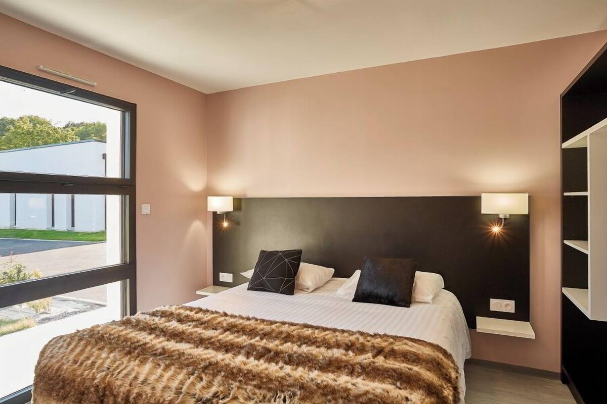 Une chambre moderne avec un grand lit, deux lampes de chevet et une vue sur un immeuble par une grande fenêtre.