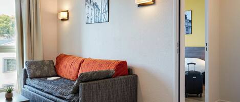 A modern hotel room with a grey sofa, a small table, and a lamp. There is a painting on the wall and a suitcase in the corner.