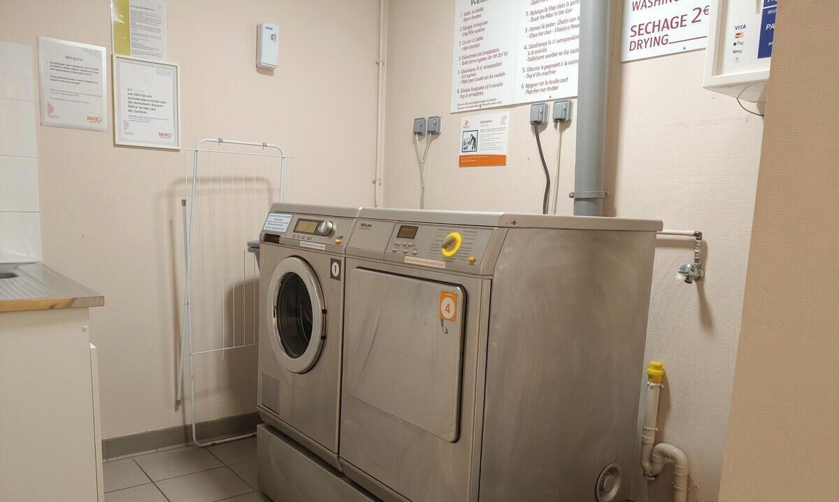 A compact laundry facility with a front-loading washing machine, a drying machine, and a wall-mounted drying rack. Instructions and a payment sign are visible.