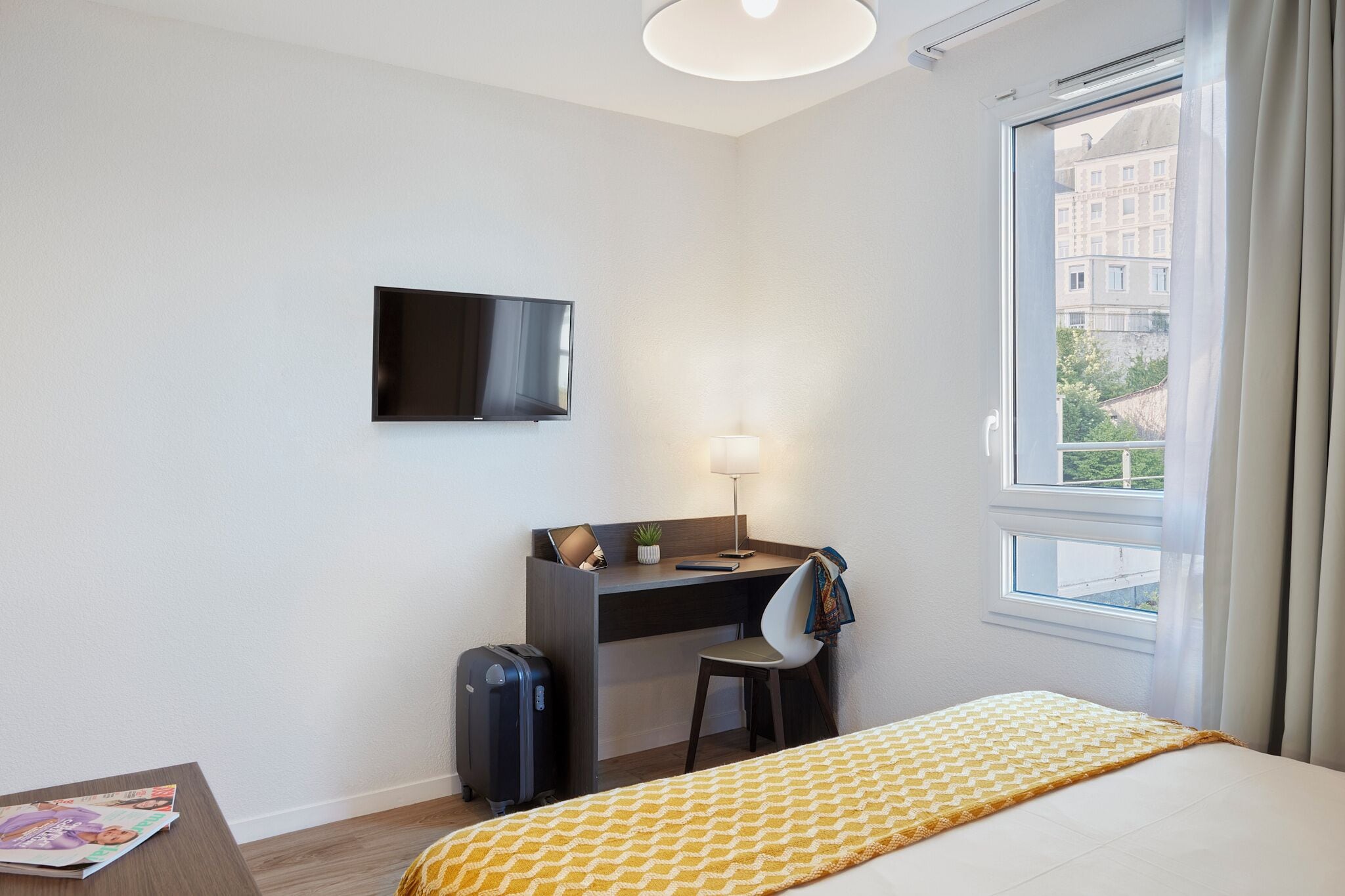 Ein Hotelzimmer mit einem Bett, einem Schreibtisch mit Stuhl, einem Fernseher, einem Koffer und einem Fenster mit Vorhängen.