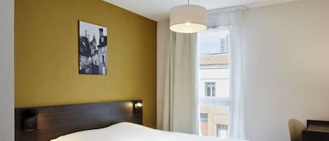 A hotel room with a bed, a bedside table with a book, a wall-mounted picture, a window with curtains, and a desk with a chair.
