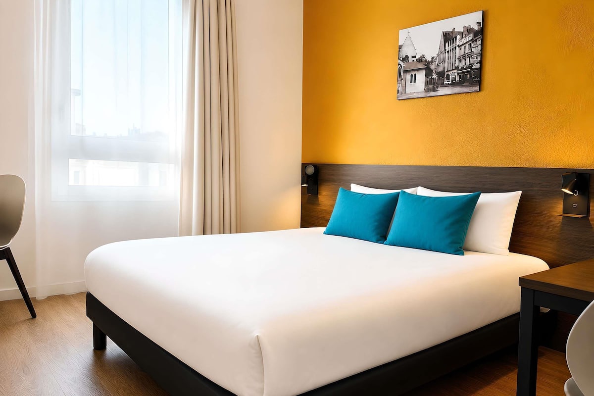 Ein Hotelzimmer mit einem großen Bett, einem Nachttisch, einem Sessel, einem Fenster mit Vorhängen und einem an der Wand hängenden Bild.