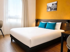 Una habitación de hotel con una cama grande, una mesita de noche, una silla, una ventana con cortinas y un cuadro colgado en la pared.