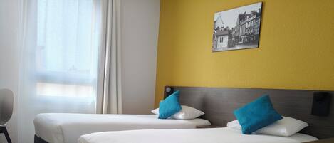 A hotel room with two beds, a wooden floor, a window with curtains, and a wall-mounted picture.