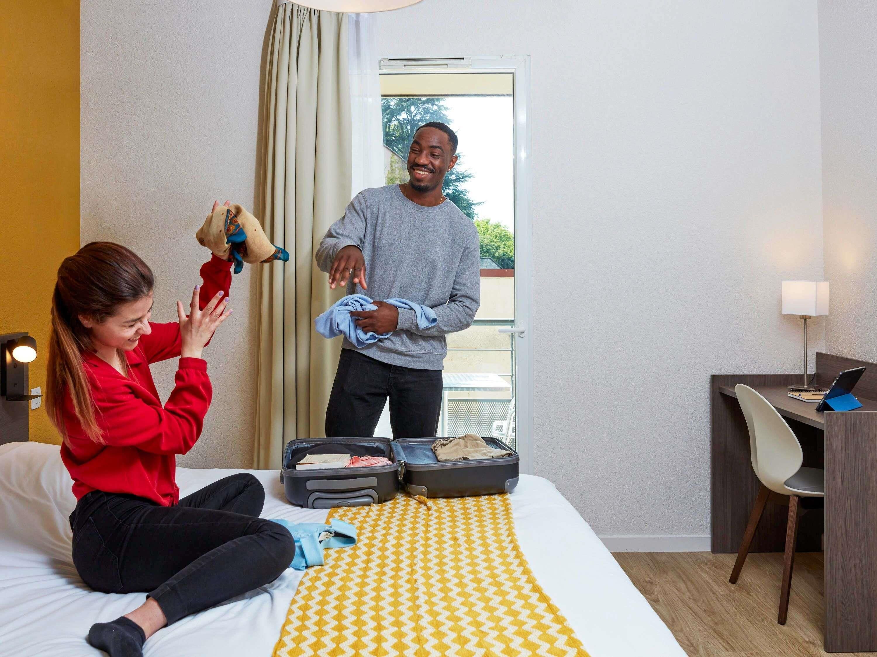 A hotel room with a bed, a desk, a chair, and a suitcase on the bed.