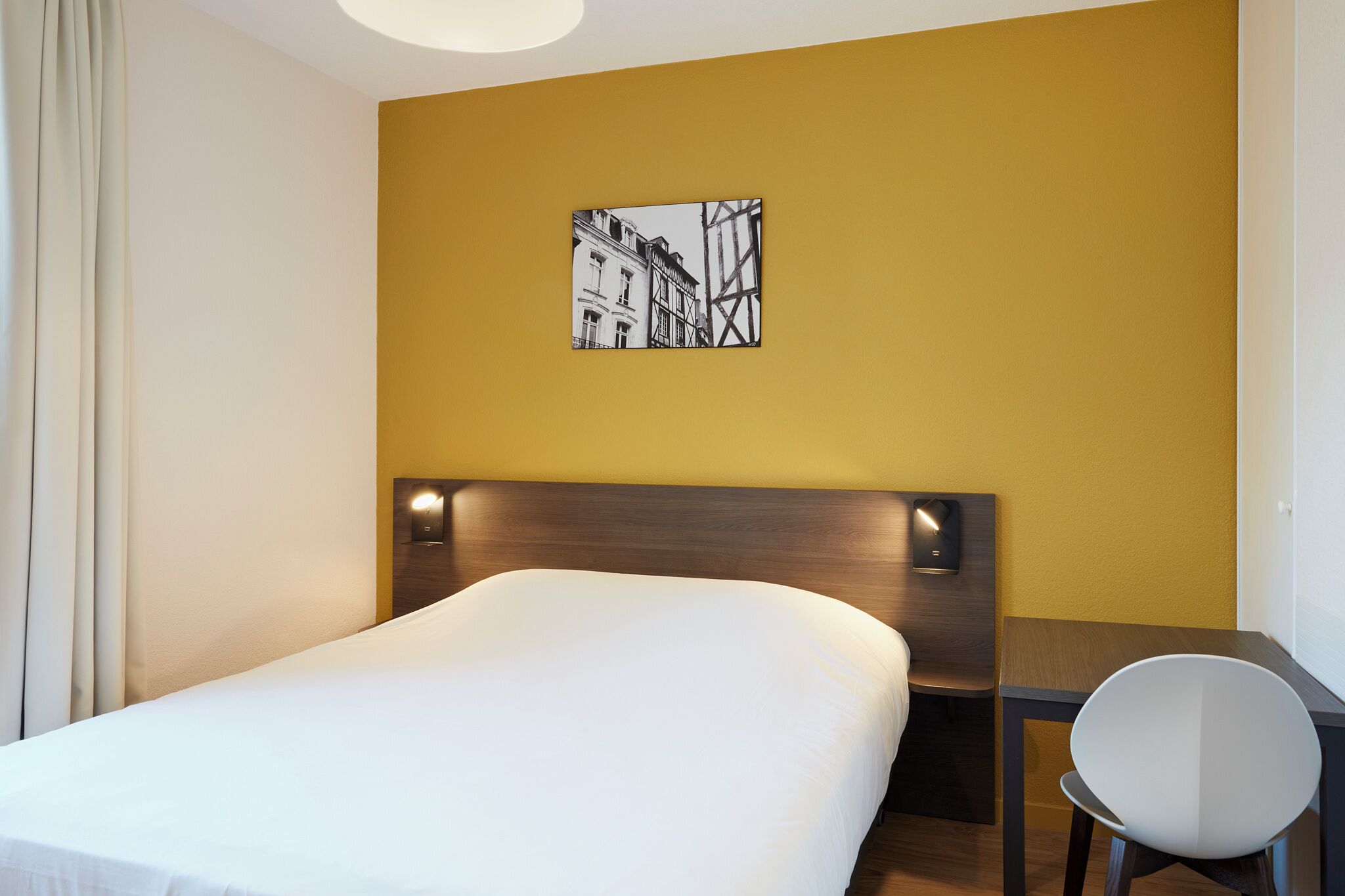 A hotel room with a bed, a desk, a chair, and a wall-mounted picture.