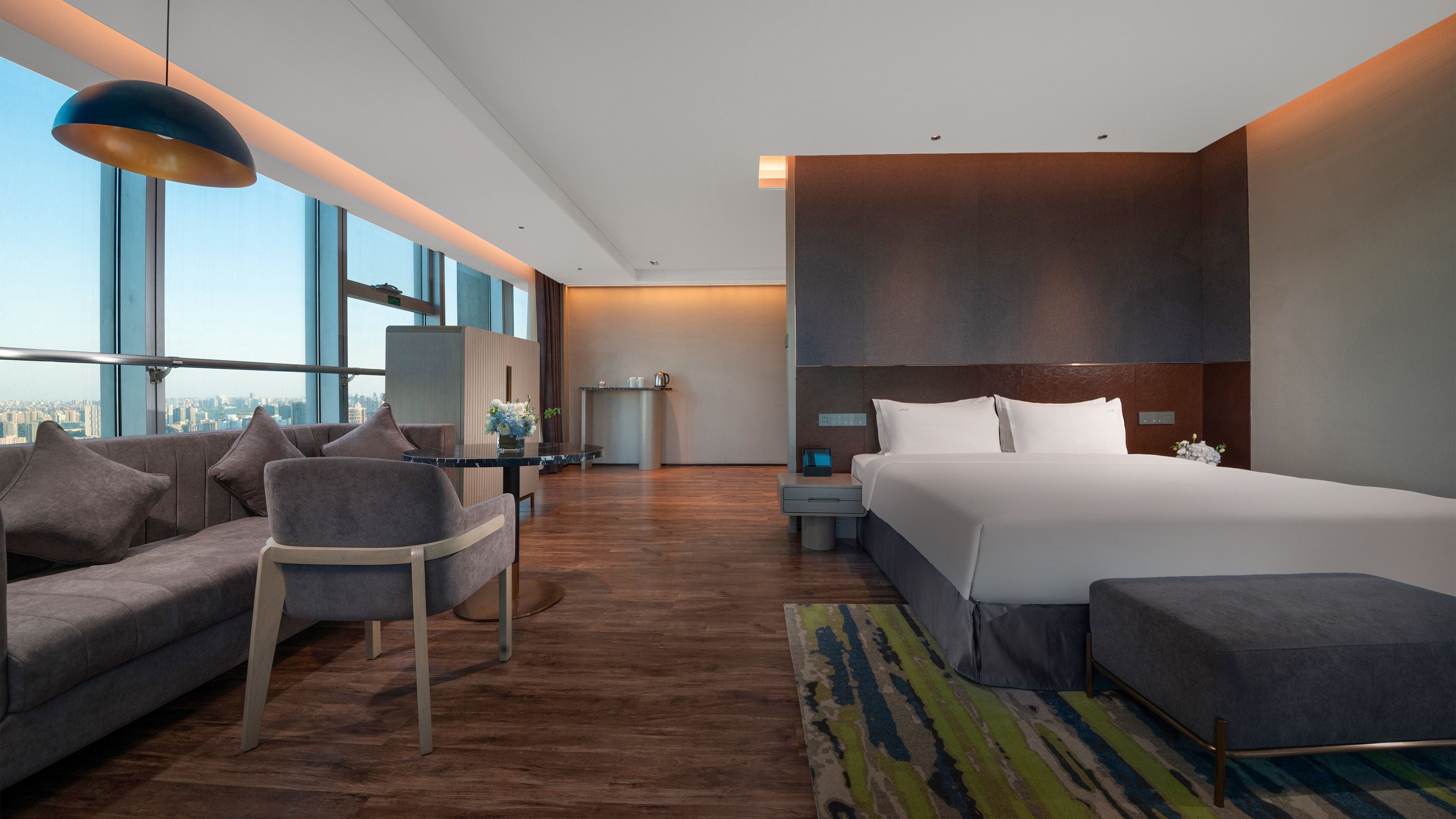 A modern hotel room with a large bed, a sofa, a desk, and a view of the city.