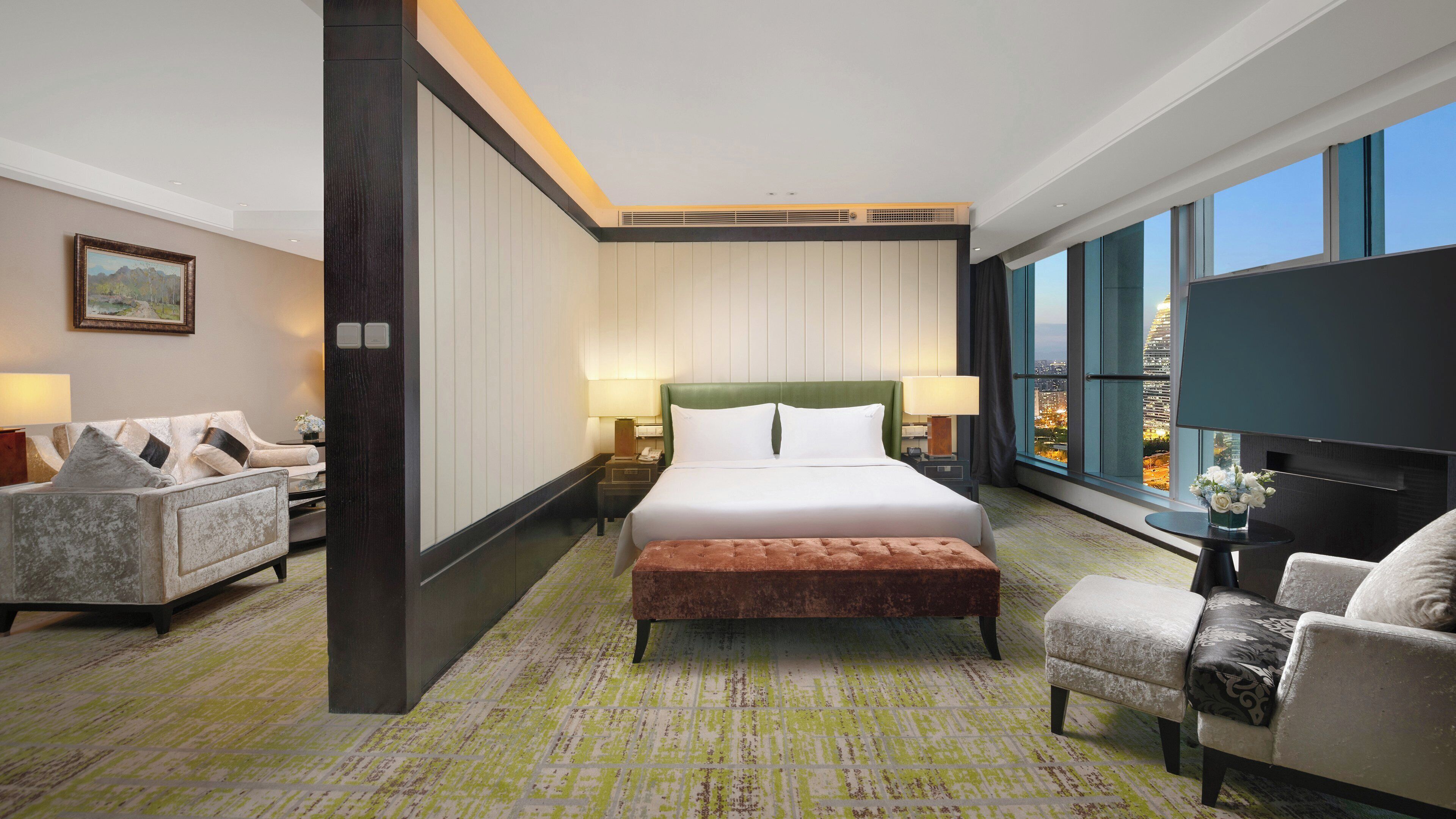 A hotel room with a large bed, two armchairs, a small table, and a view of the city through large windows.