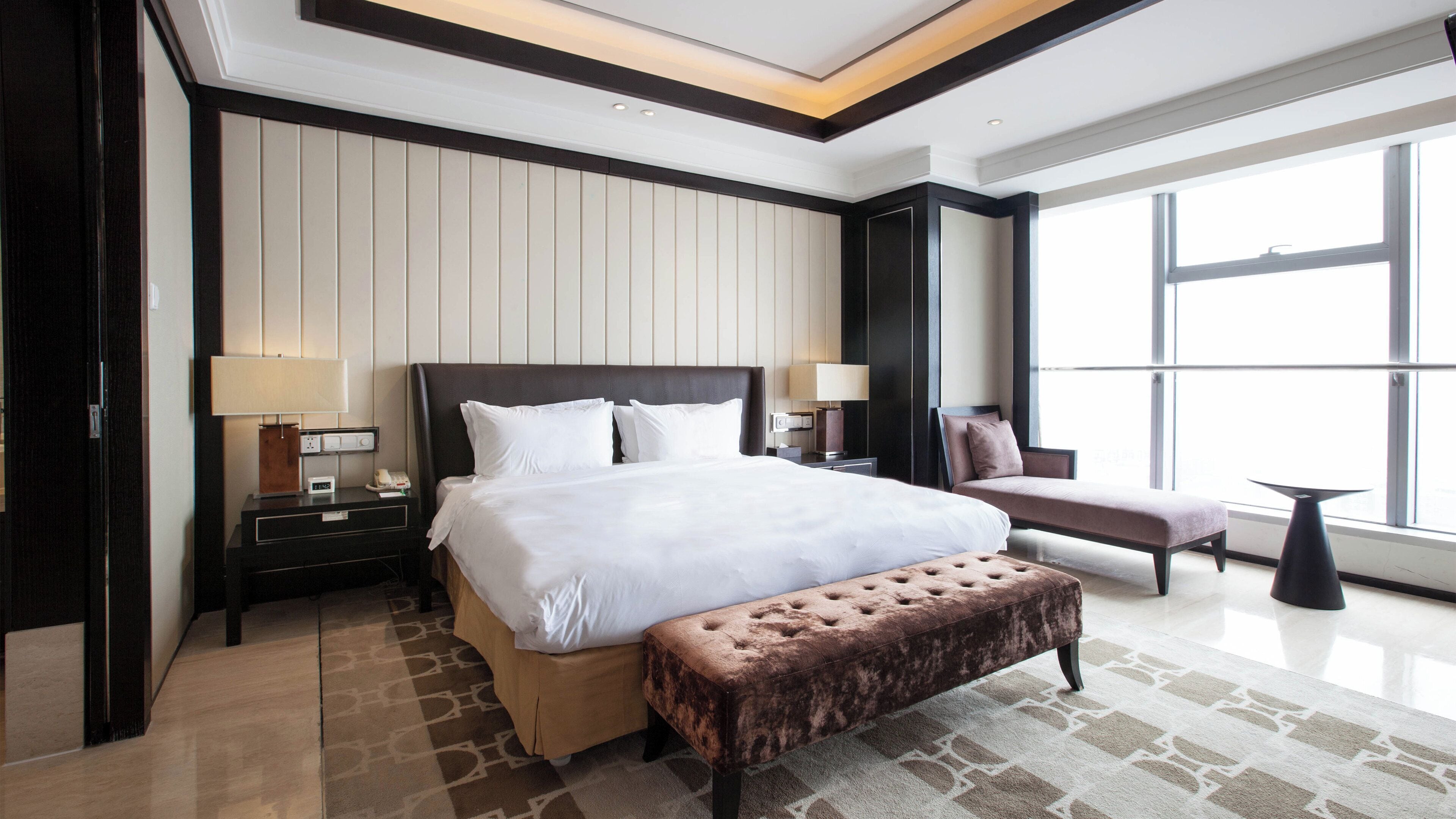 A hotel room with a large bed, bedside tables, a bench, and a window.
