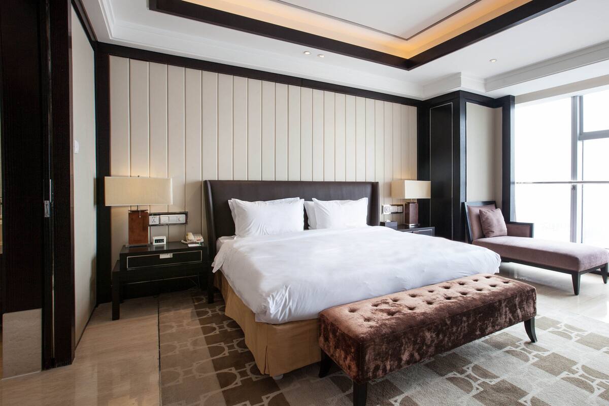 A modern hotel room with a large bed, a bedside table with a lamp, a chair, and a window providing a view of the outside.