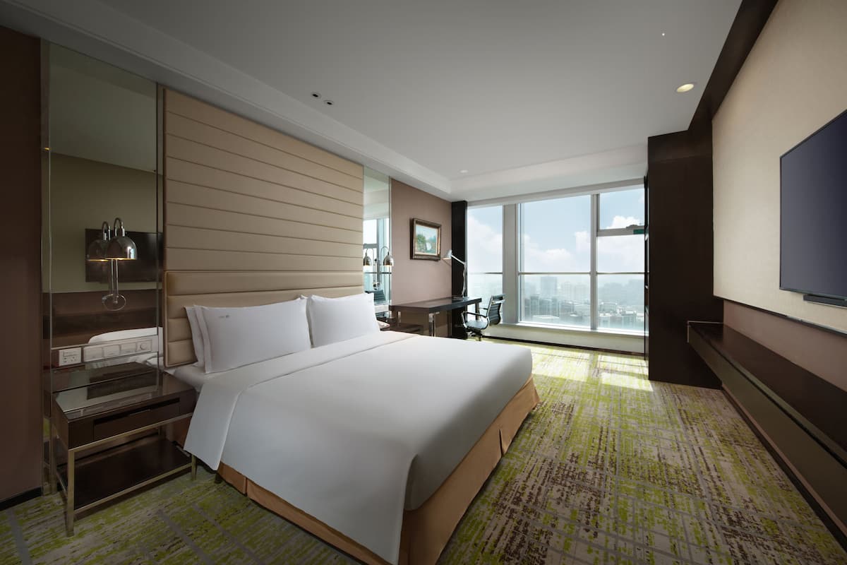 A modern hotel room with a large bed, a desk, a chair, a flat-screen TV, and a large window offering a city view.