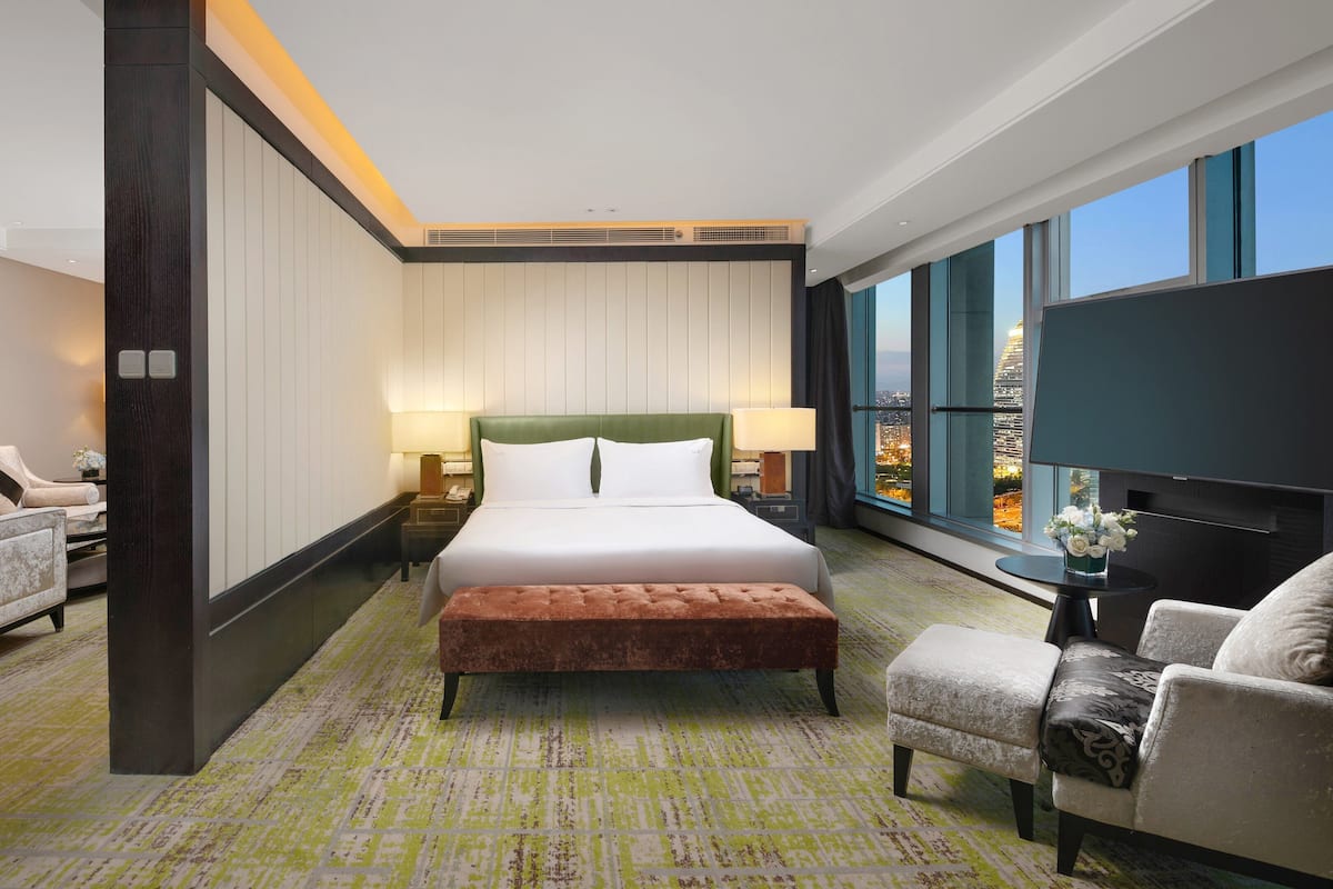 A hotel room with a large bed, two armchairs, a small table, and a large window offering a city view.