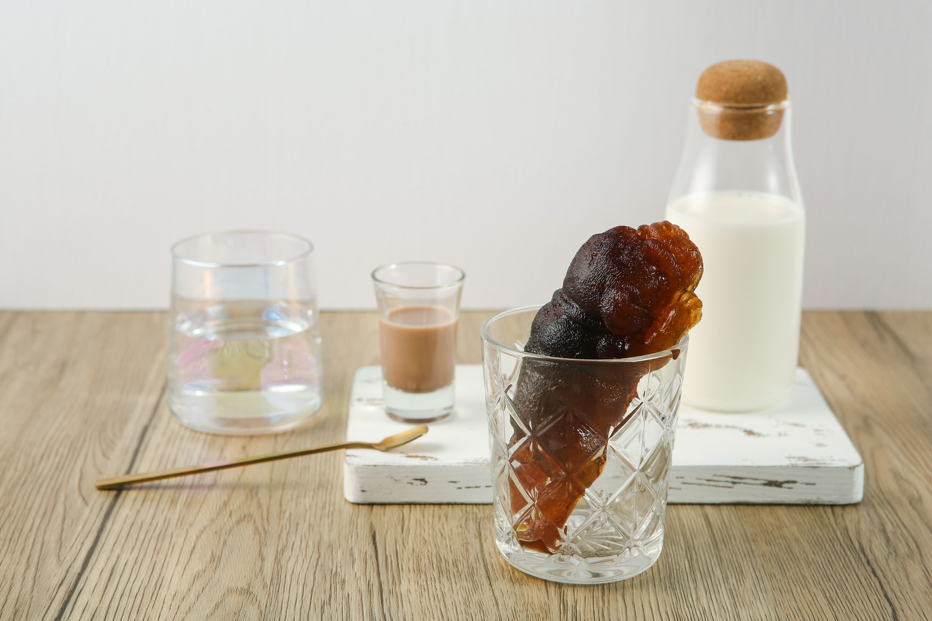 Un vaso con un líquido oscuro, un vaso pequeño con un líquido de color claro y una botella de leche sobre una mesa de madera.