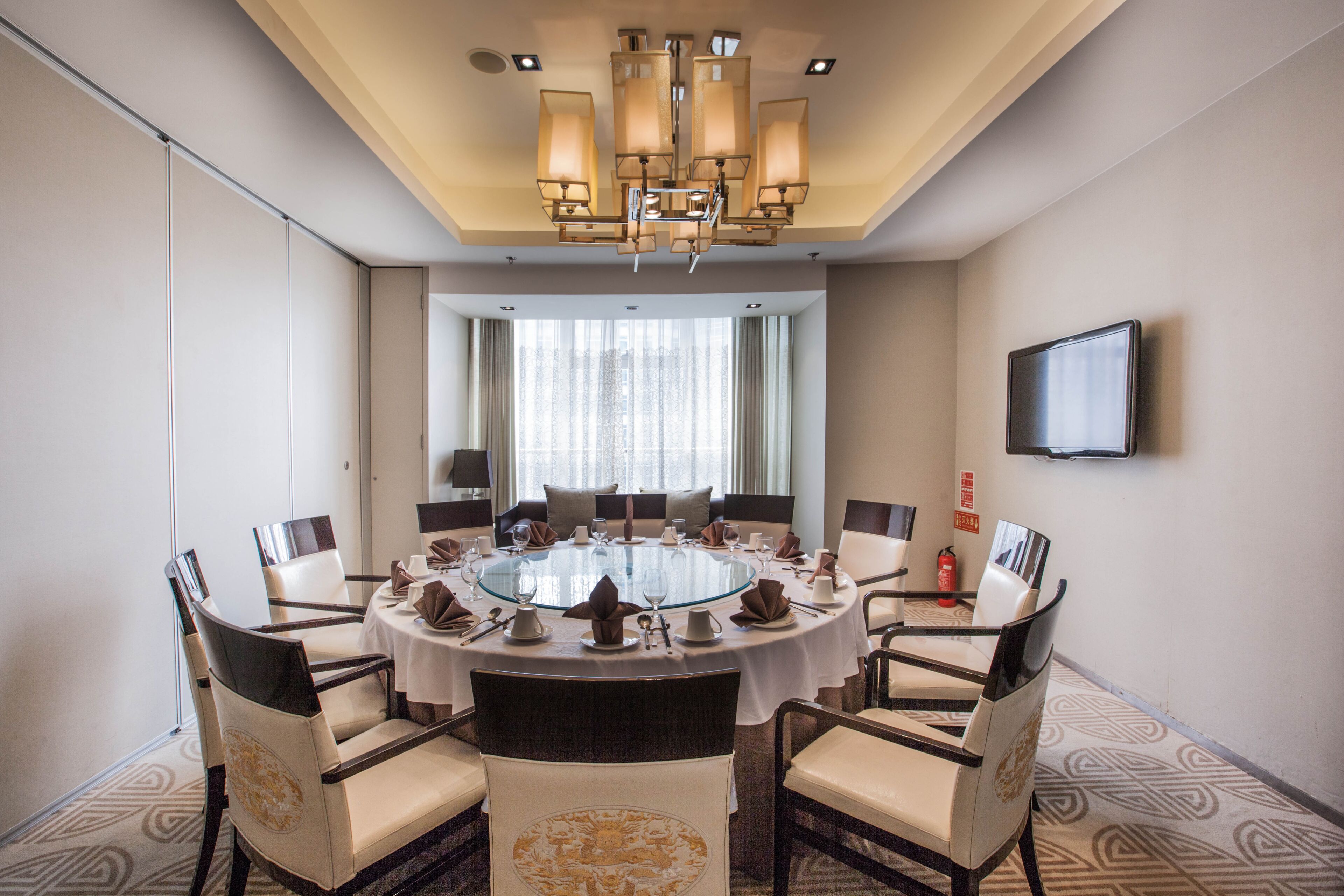 A conference room with a round table, eight chairs, a chandelier, a television on the wall, and a window with curtains.