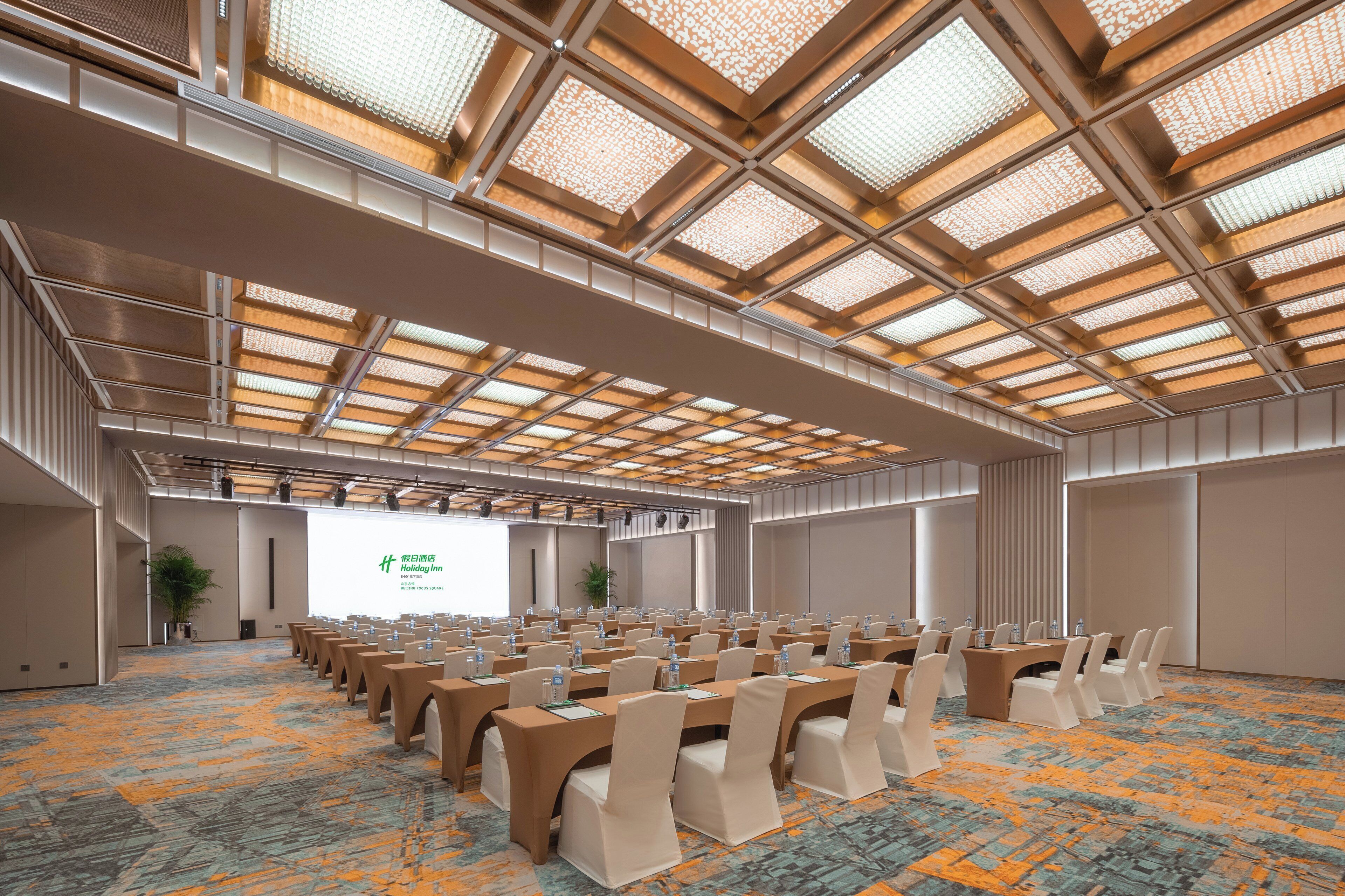 A conference room with a high ceiling, patterned ceiling panels, and rows of tables with chairs.