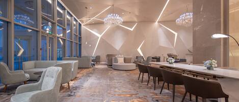 A modern hotel lobby with a large seating area, a long table, and a patterned carpet.
