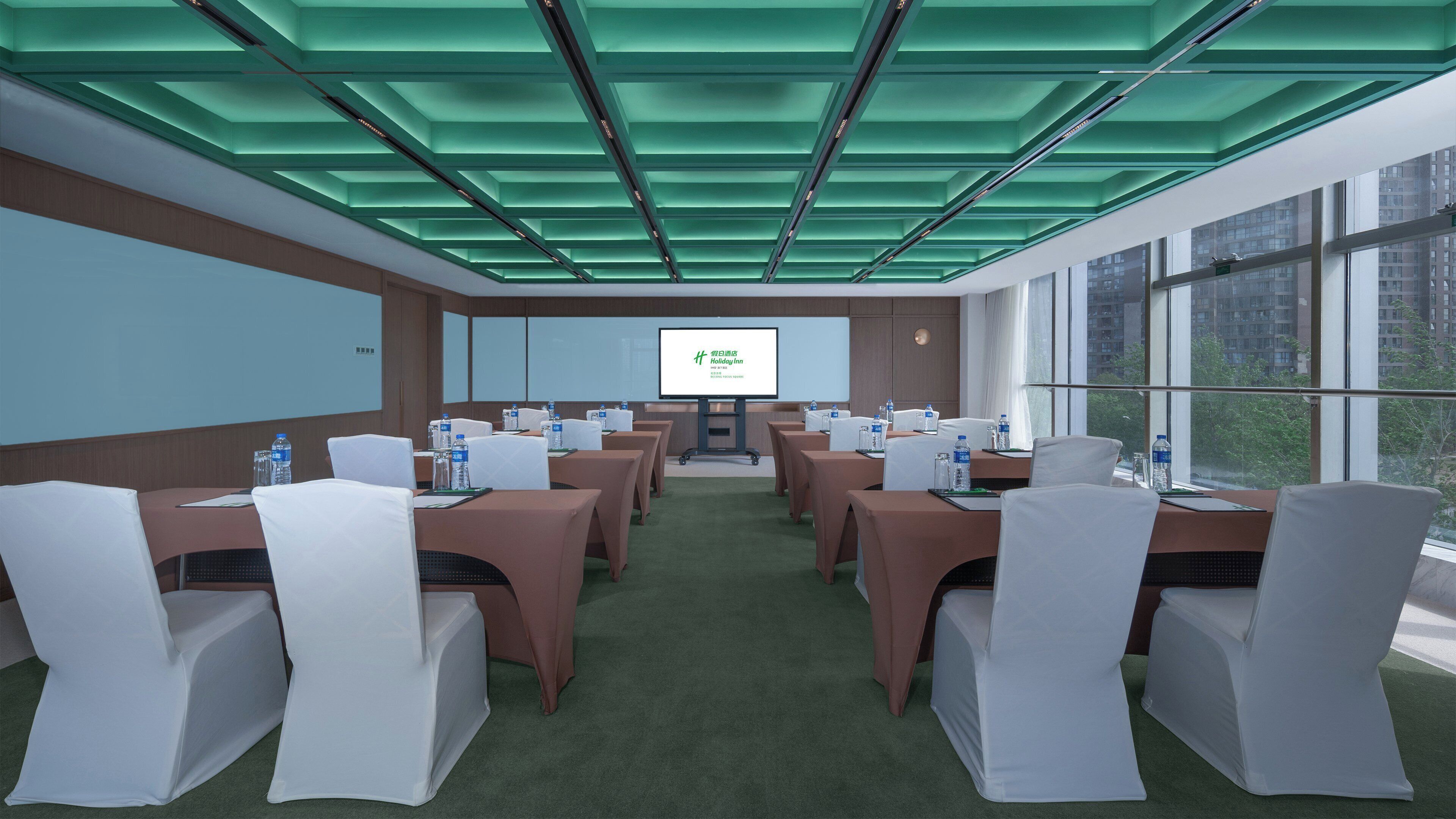 Ein Konferenzraum mit grüner Decke, mehreren Tischen mit Stühlen, einer Projektionsleinwand und Blick auf Gebäude durch große Fenster.