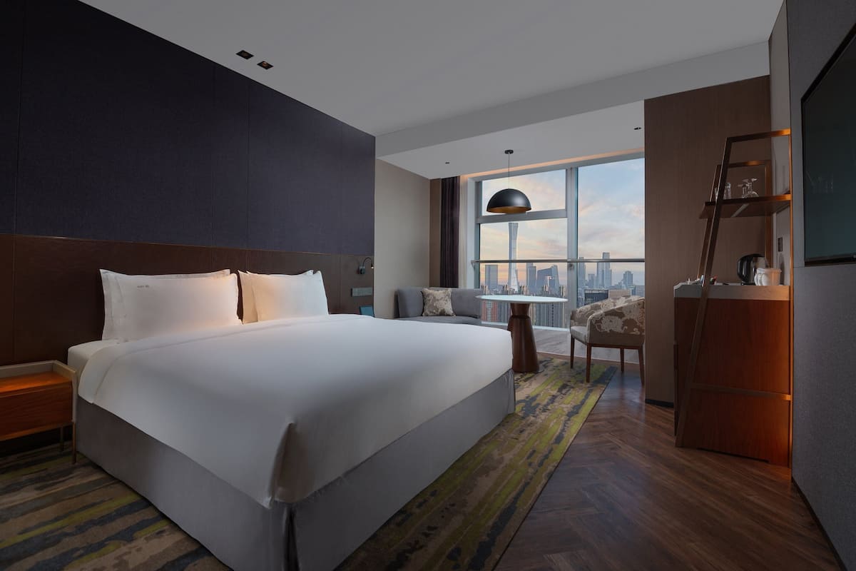A modern hotel room with a large bed, a city view, a desk, and a chair.