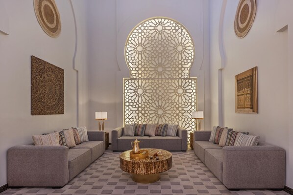 Un salon moderne avec un sol à carreaux, deux canapés gris ornés de coussins à motifs, une table basse ronde agrémentée d’objets décoratifs et un grand paravent décoratif aux motifs complexes.