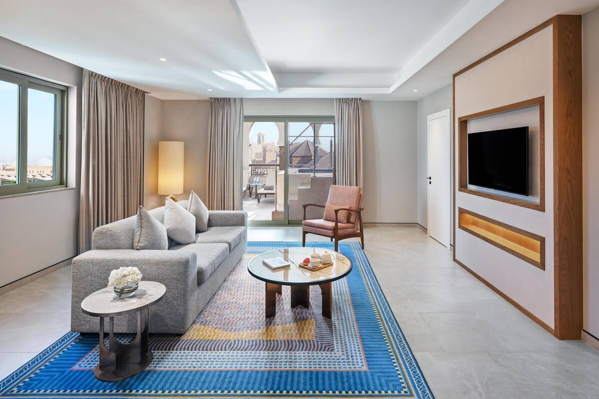 Ein modernes Hotelzimmer mit grauem Sofa, einem runden Couchtisch, einem an der Wand montierten Fernseher und Blick auf die Stadt durch große Fenster.