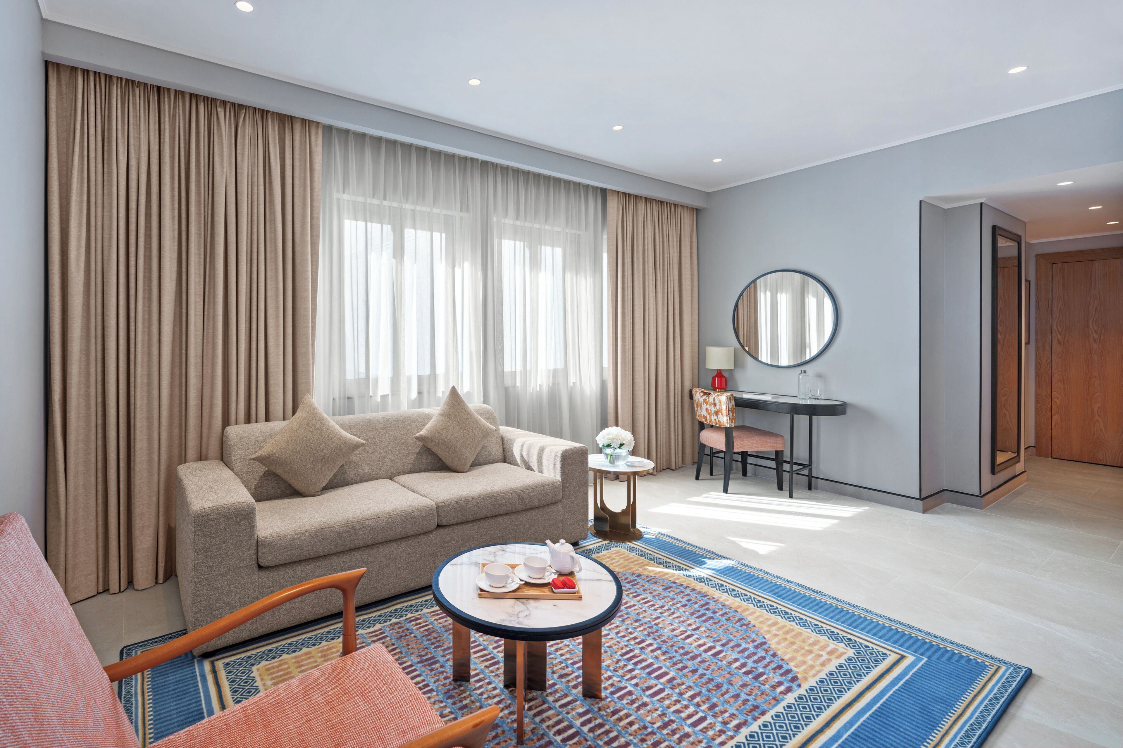 Un salon moderne avec un canapé, une petite table ronde et un fauteuil. On y trouve également un bureau avec une chaise et un miroir. La pièce est dotée d’une grande fenêtre avec des rideaux.