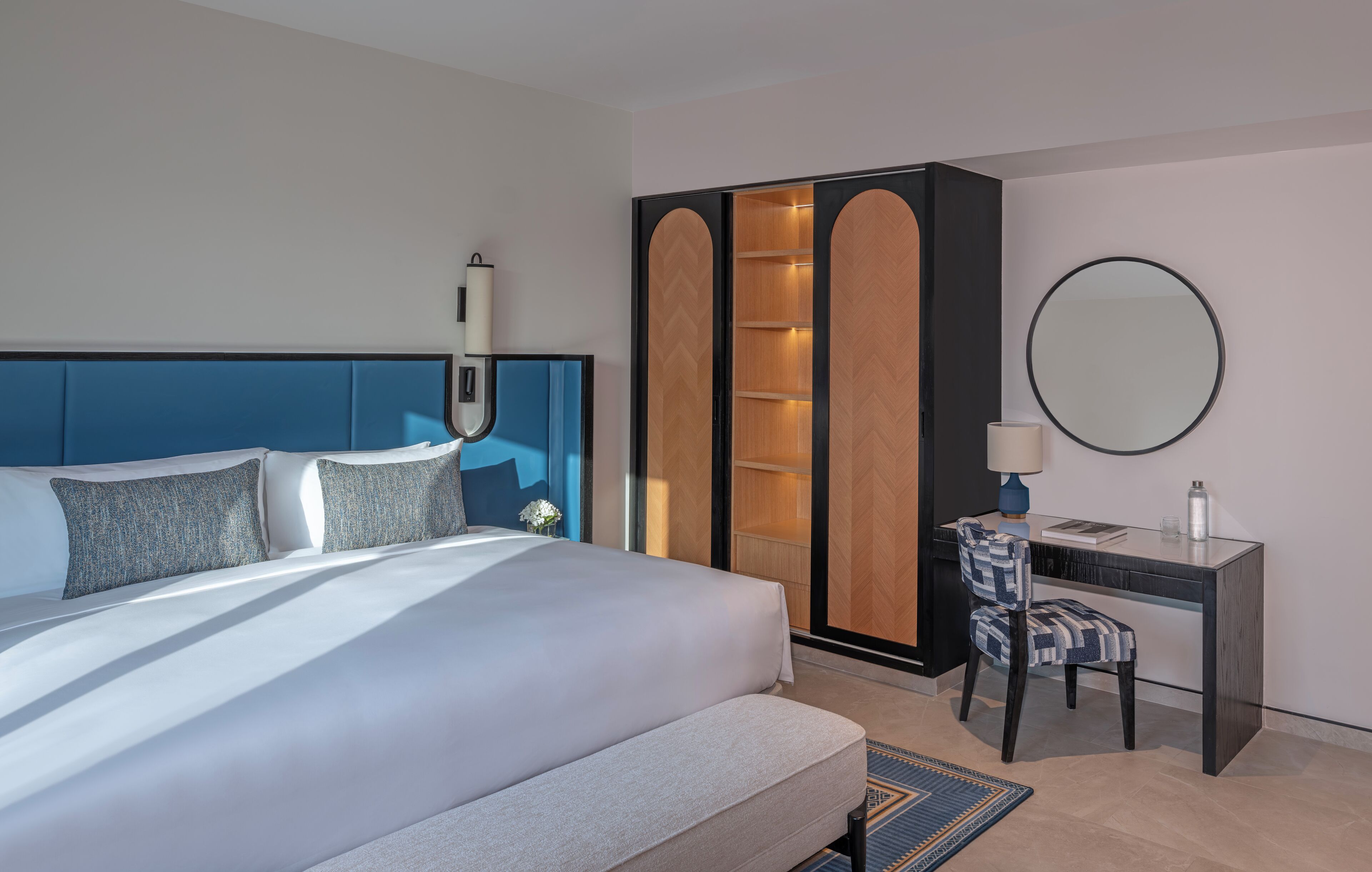 Une chambre d’hôtel moderne avec un grand lit, un bureau avec une chaise, une armoire et un miroir rond.