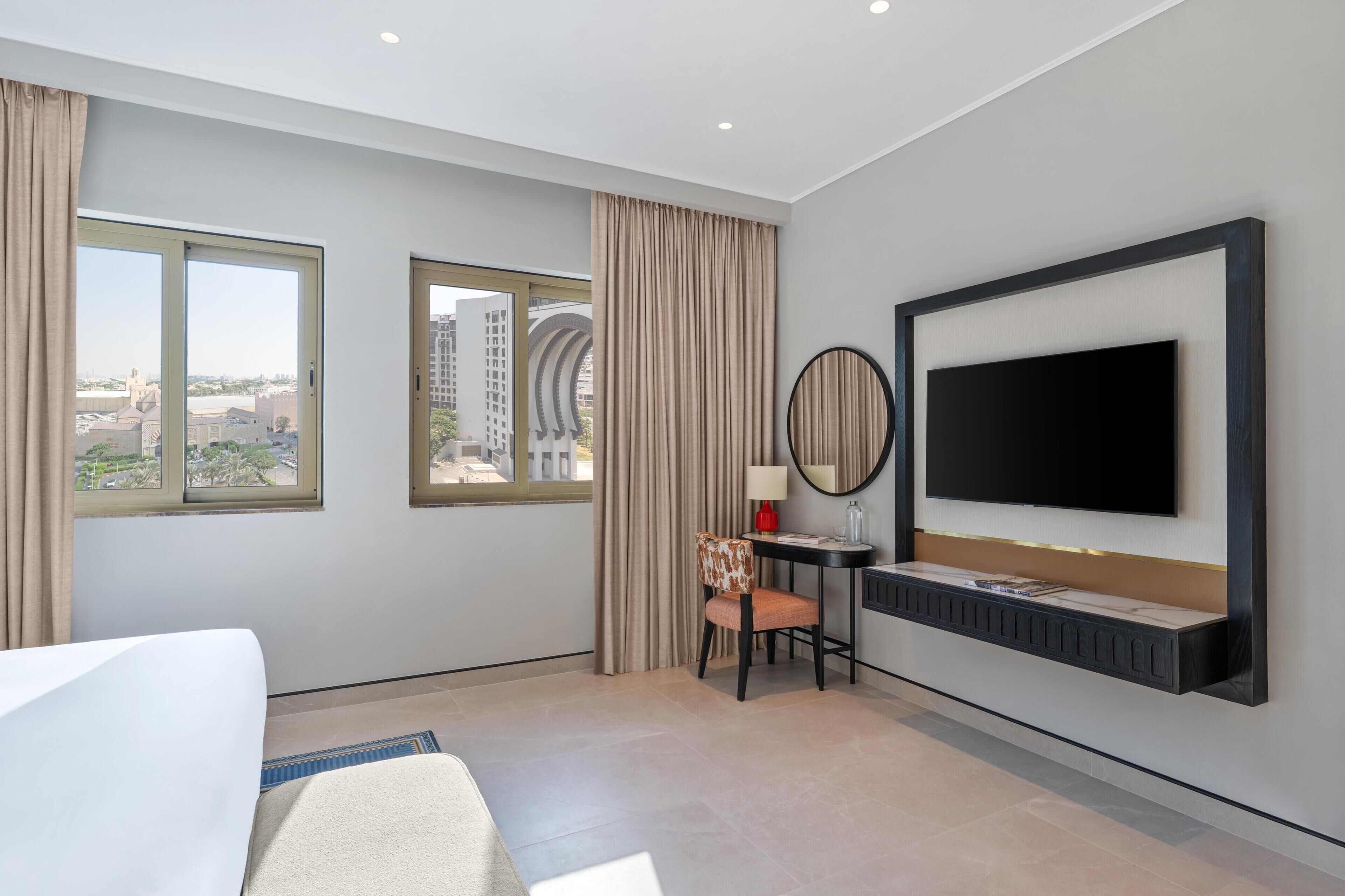 A hotel room with a bed, a desk with a chair, a TV mounted on the wall, and two windows with curtains.