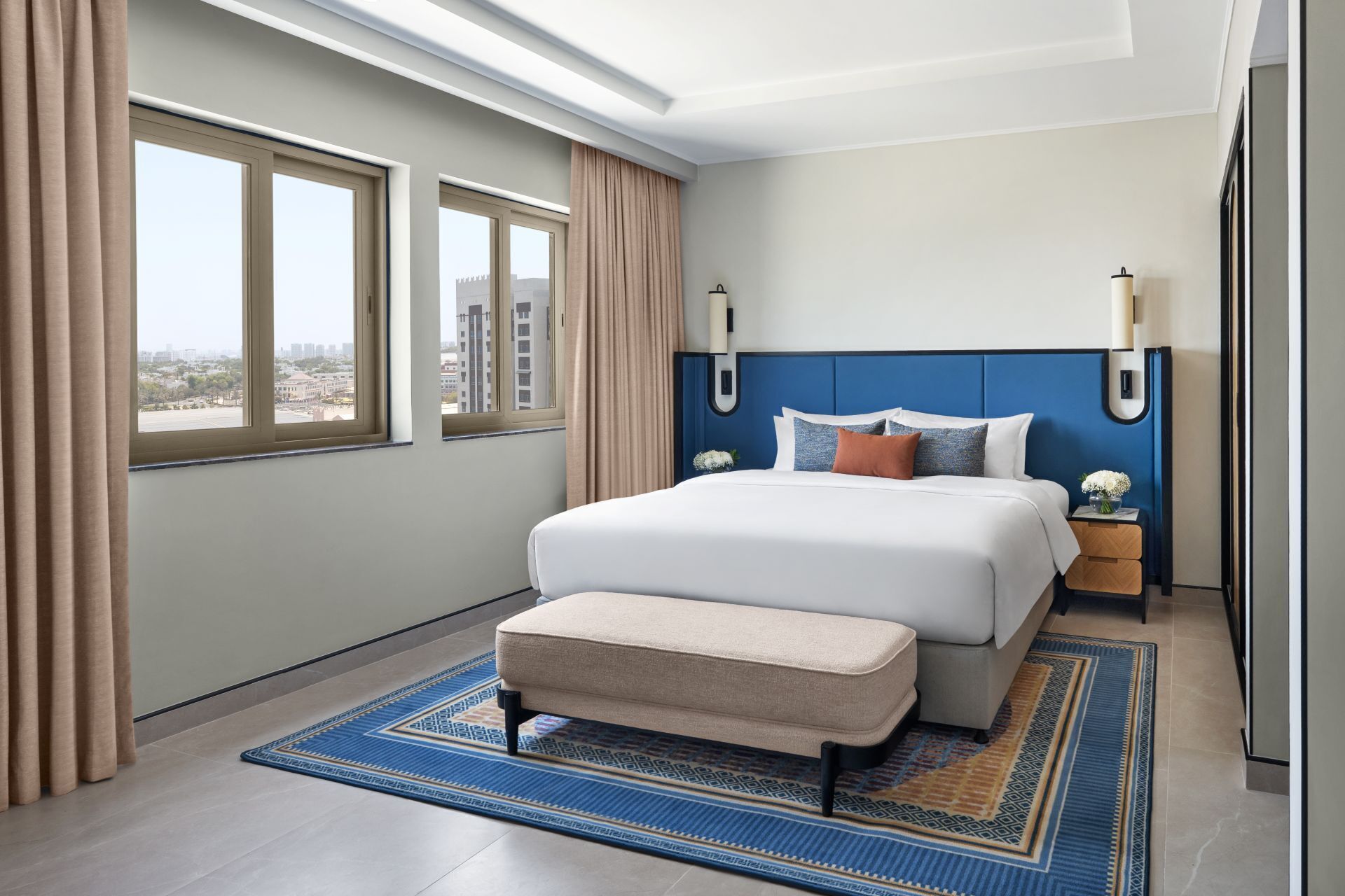 Une chambre d’hôtel moderne avec un grand lit, deux tables de chevet, un banc et une vue sur la ville.